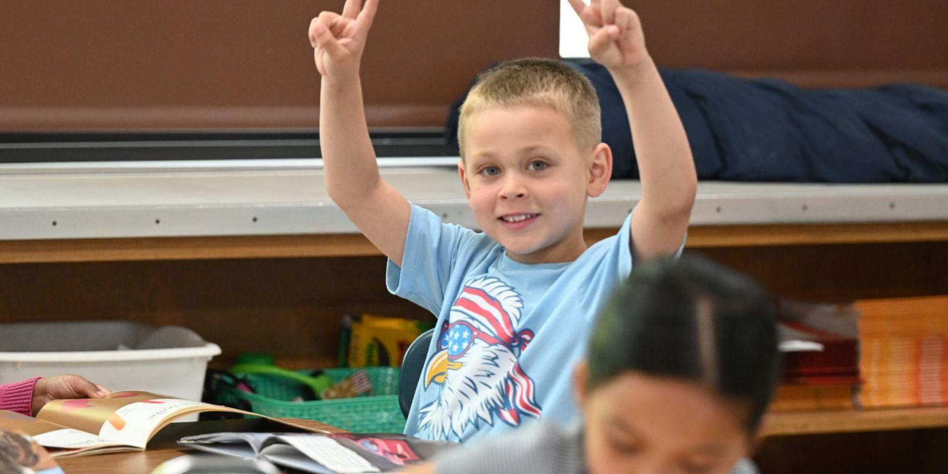 Boy raising peace signs with a smile in a classroom setting.