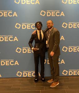 Image of a student and his advisor standing in front of a backdrop that says DECA