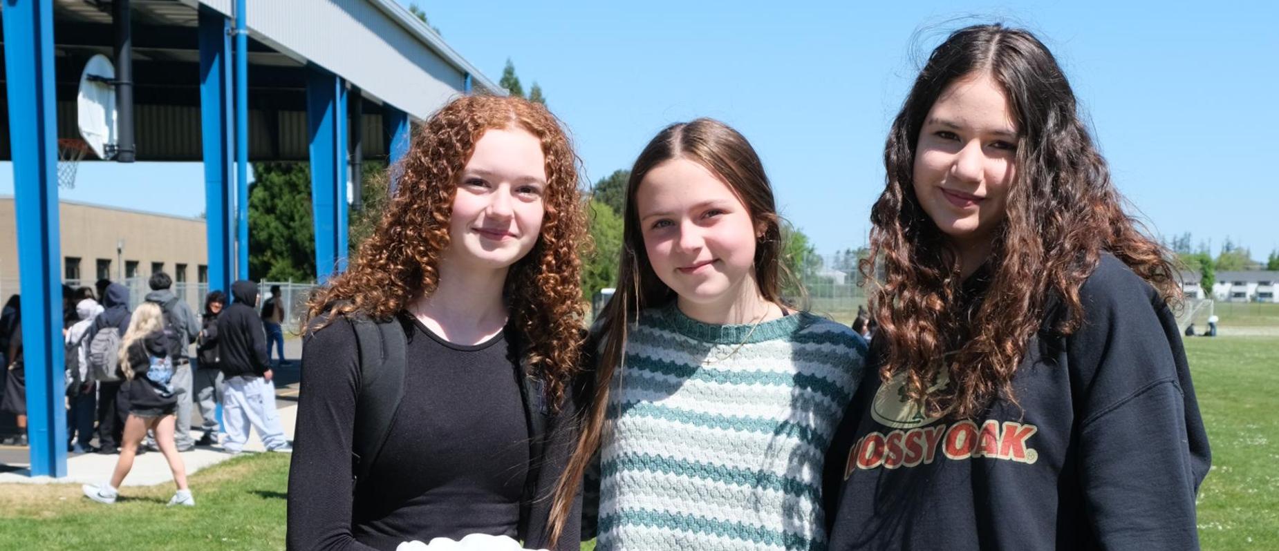 Three girls posing together outdoors, smiling, with a green field in the background.