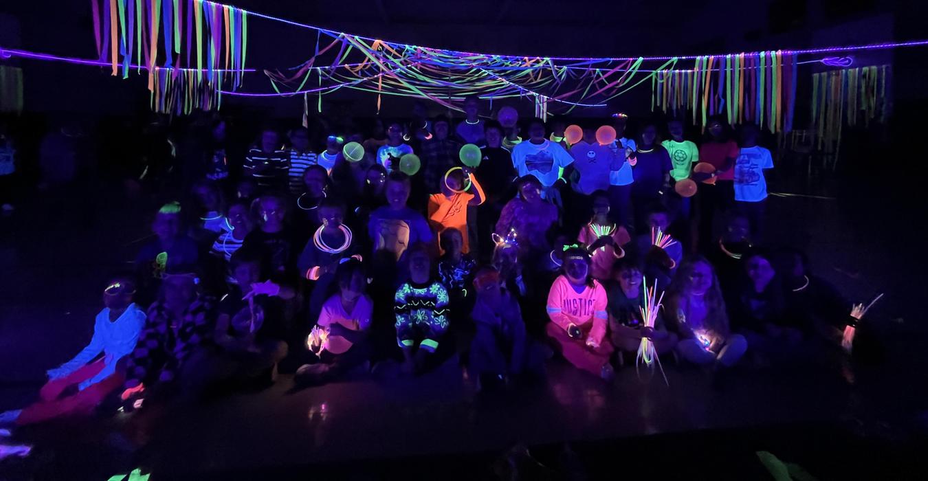 A crowd of people in a dark room wearing glow sticks and surrounded by colorful neon decorations.