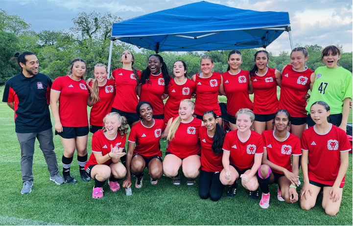 Girls Varsity Soccer players stand in red uniforms and black shorts  next to coach outside on a cloudy day