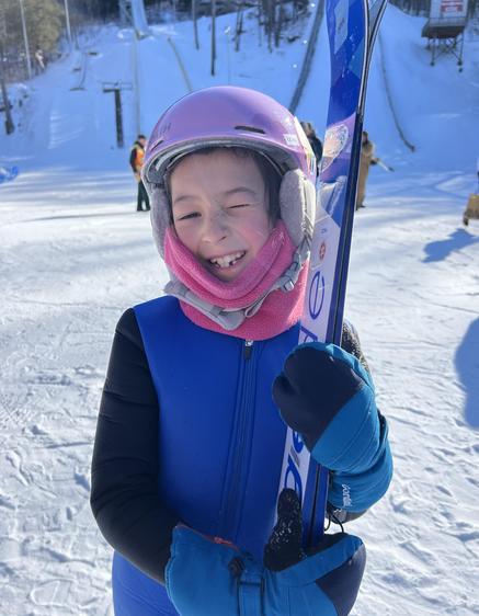 Child in ski gear smiles while holding skis on a snowy slope.