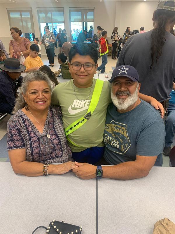 Grandson wearing green shirt and safety patrol belt standing wiht arms around grandparents