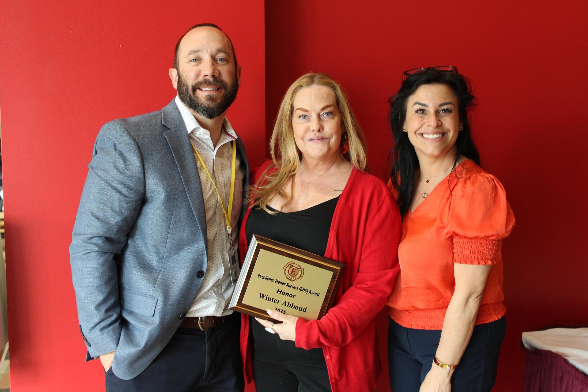 A teacher, standing in between two colleagues, holding an award certificate