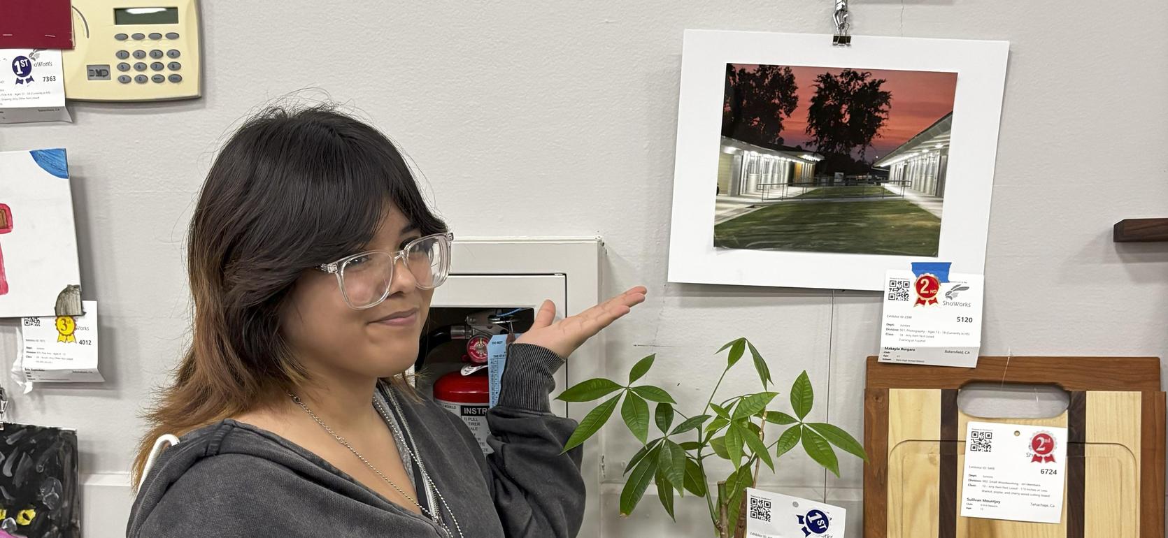 Woman in a gray hoodie points to a potted plant on display near wooden items.