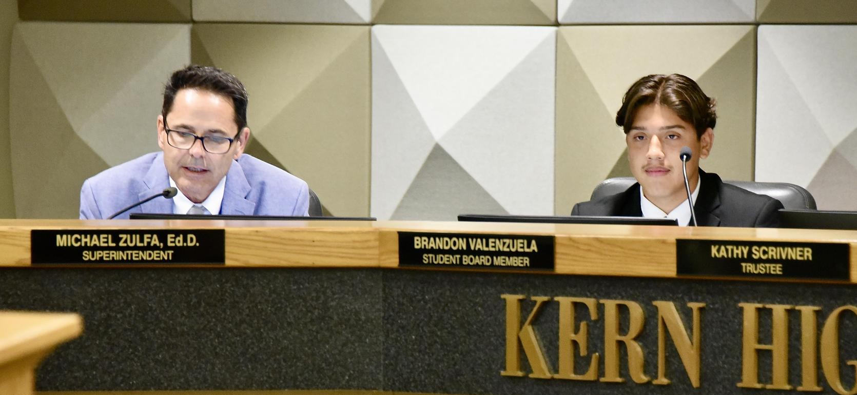 Two men seated at a board meeting table, with names displayed in front of them.