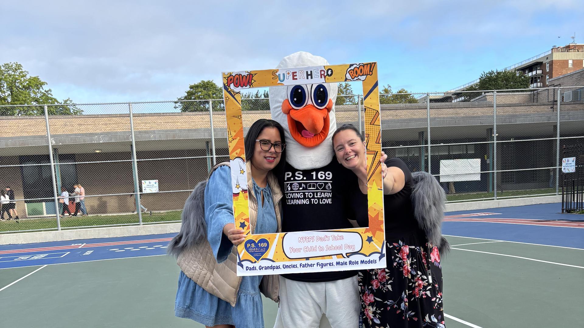 Two women in dresses posing with a large, friendly mascot in a schoolyard.