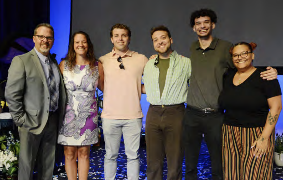 Superintendent Peter Bailey stands with his family for a group photo on stage. From left to right are Peter, his wife Justina in a purple and white patterned dress, and their children JD, Austin, Jamal, and Jasmine. Each family member is smiling and dressed in a mix of casual and semi-formal attire, showing warmth and togetherness. The background features a stage setup with flowers and a blue-lit backdrop, creating a celebratory atmosphere.