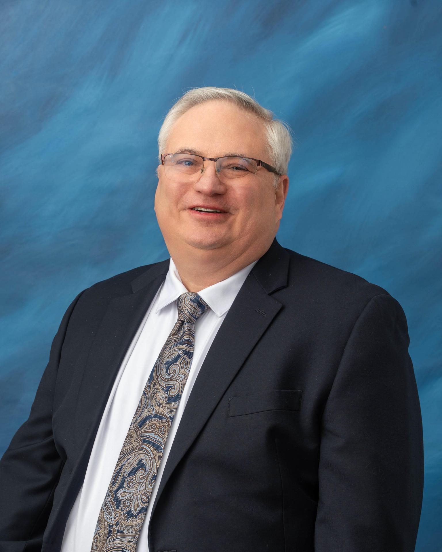  Photo of Dr. Russell Miller in a suit sitting in front of a blue background.