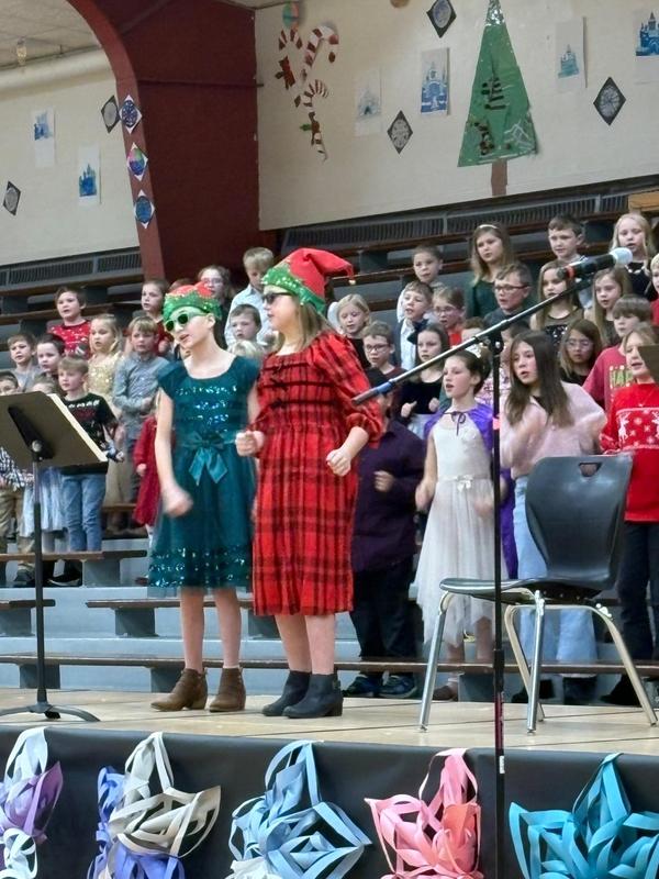 Two girls in festive outfits singing on stage with a large group of students behind them.