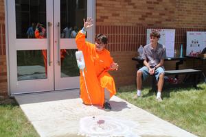student in orange poncho sits beside soda bottle rocket blasting off