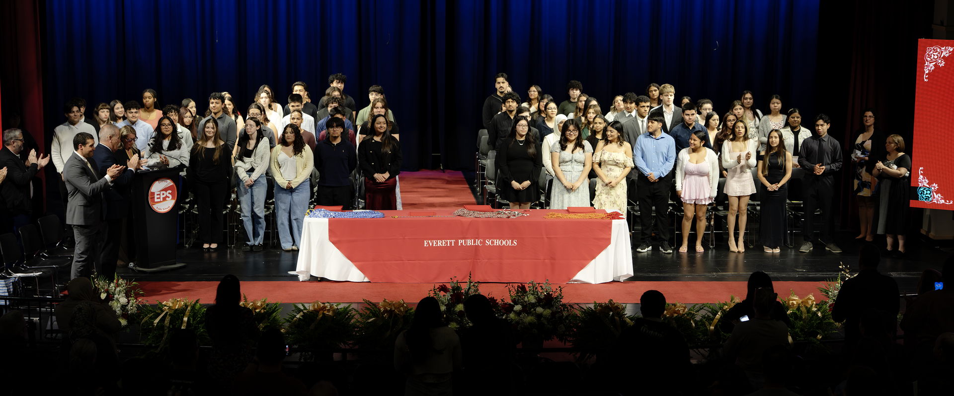 Students and faculty stand on stage during a graduation event with applause.
