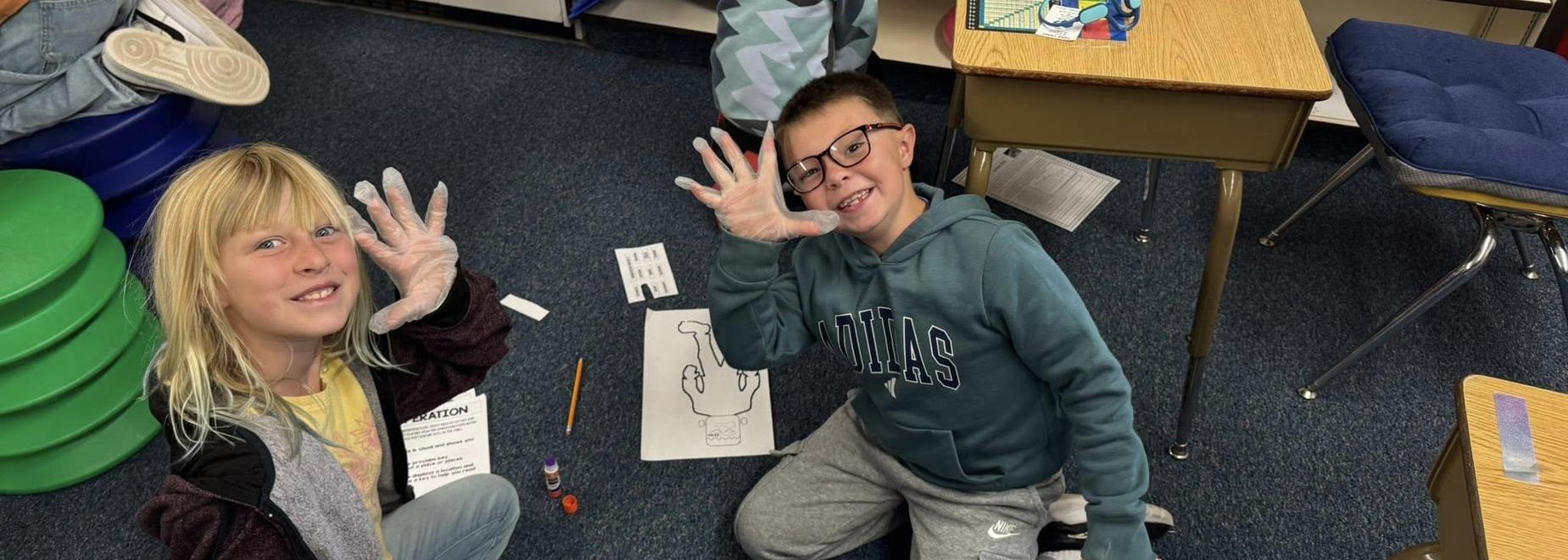 Two children in a classroom, smiling and wearing gloves, with art supplies and papers.
