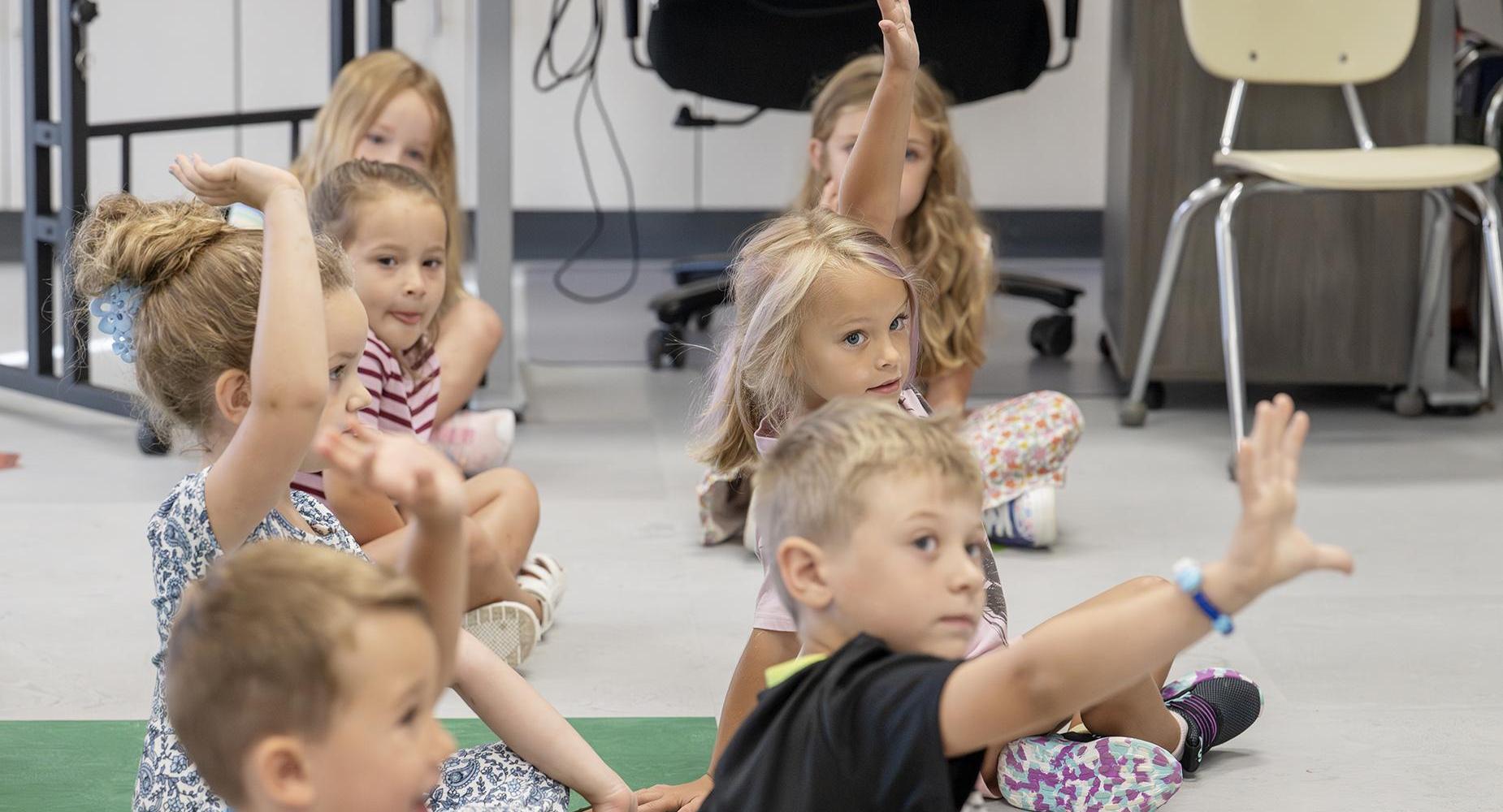 Elementary students raise hands while sitting on a carpet.