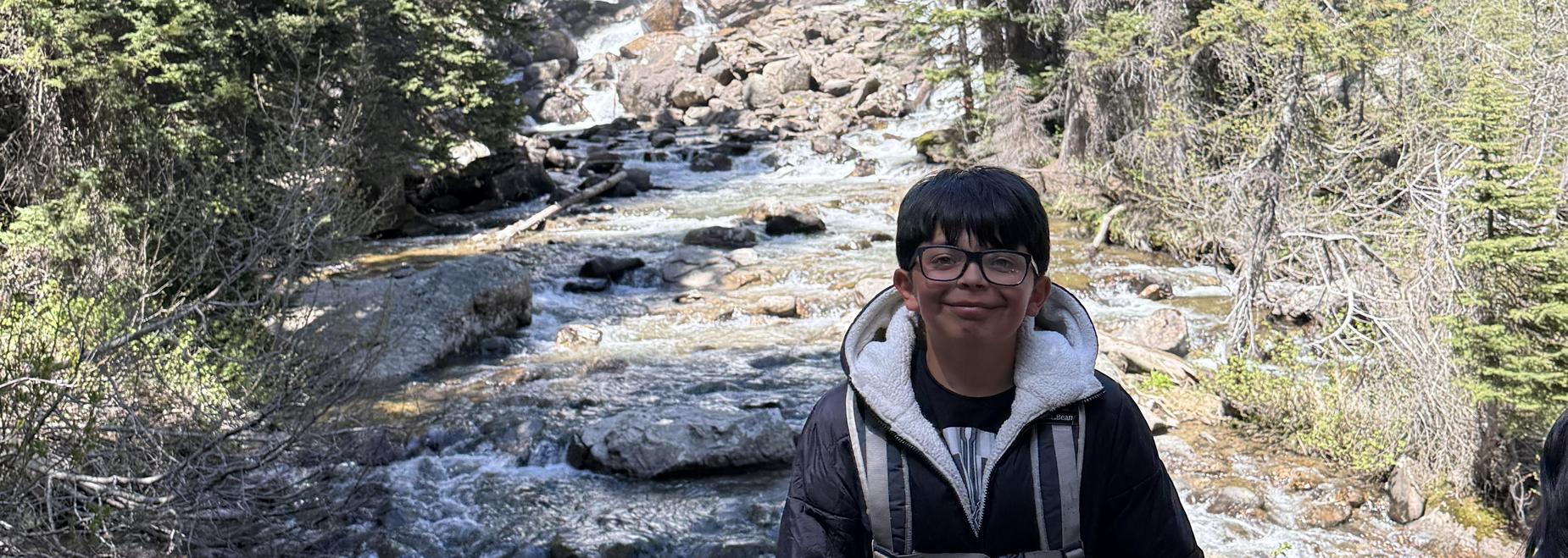 A child stands in front of a waterfall, surrounded by trees and rocky terrain.