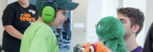 Student wearing green shirt and headphones with 2 adults with puppets