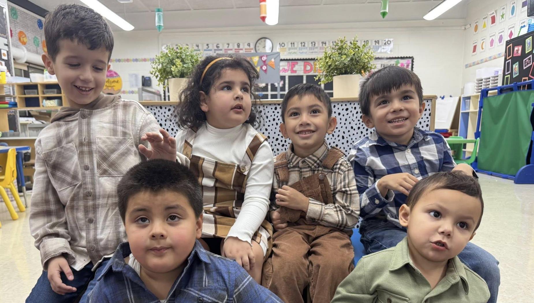 A group of six children seated on the floor, smiling in a classroom with educational displays.