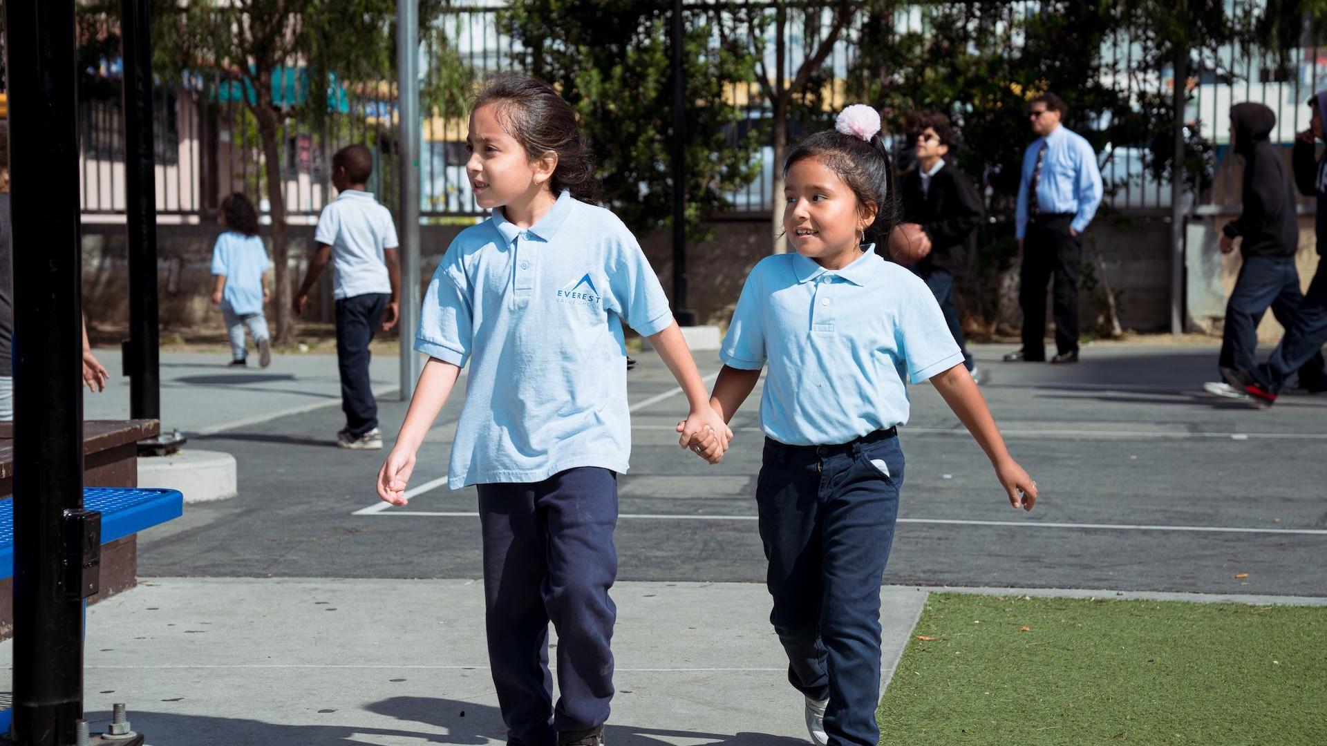 Two girls holding hands while walking on a playground.