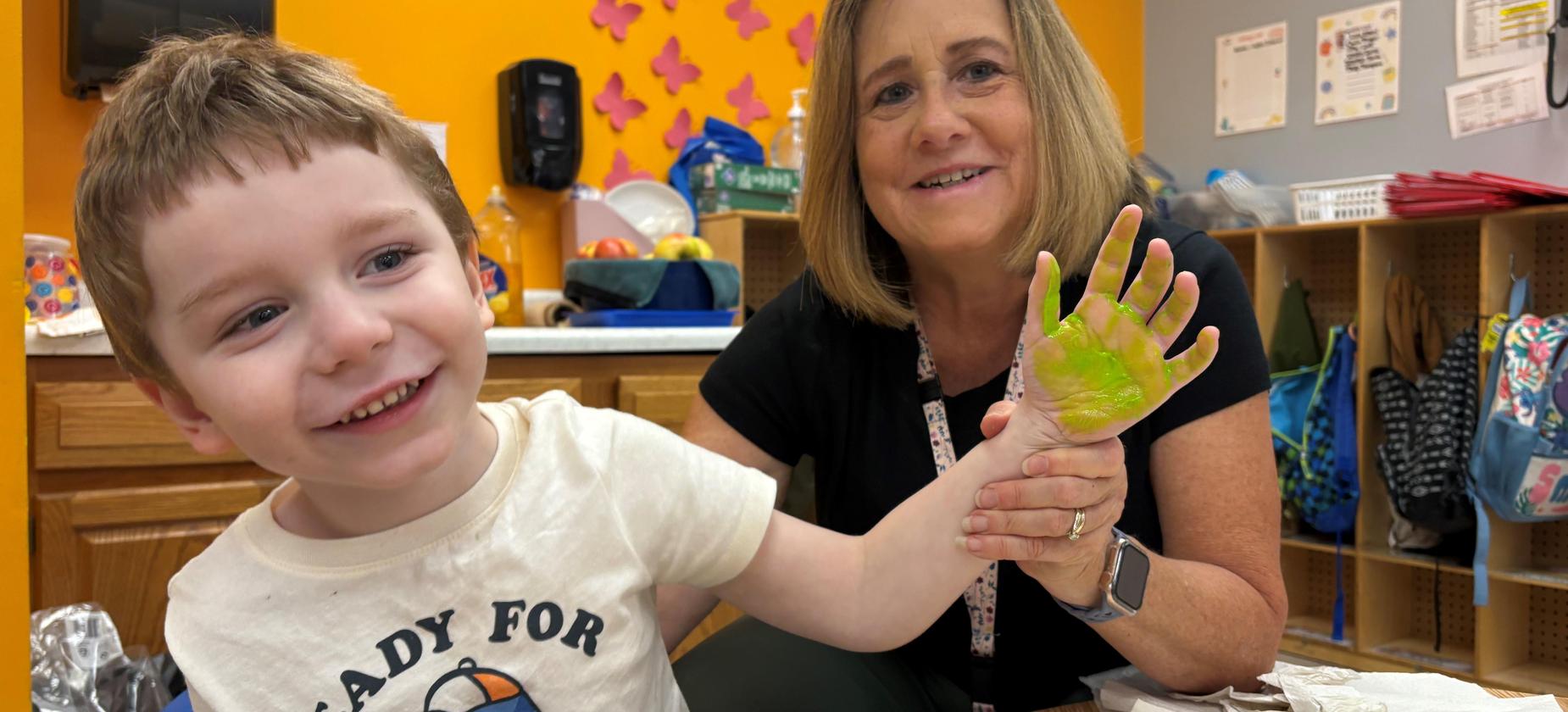 Boy smiling while showing his painted hand to a teacher in a colorful classroom.