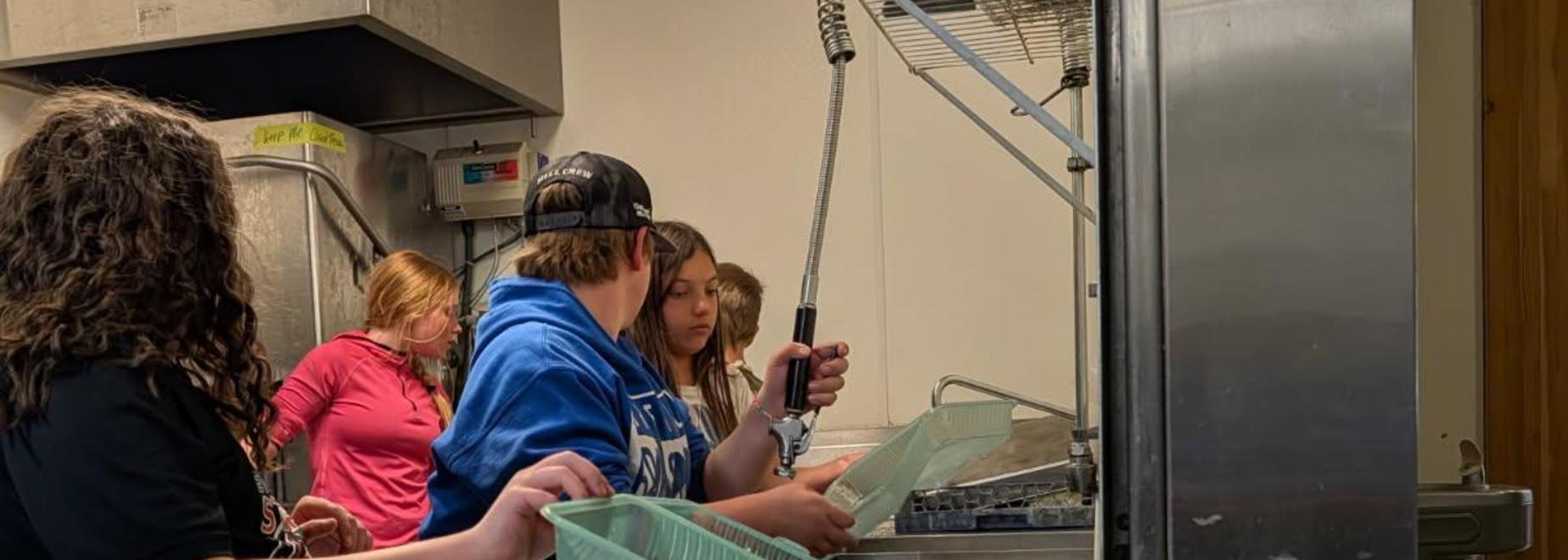 Teens washing dishes in a kitchen with green baskets.