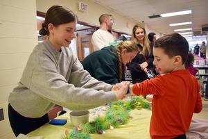 Girl assists a young boy in making a craft at a table with decorations and adults around.
