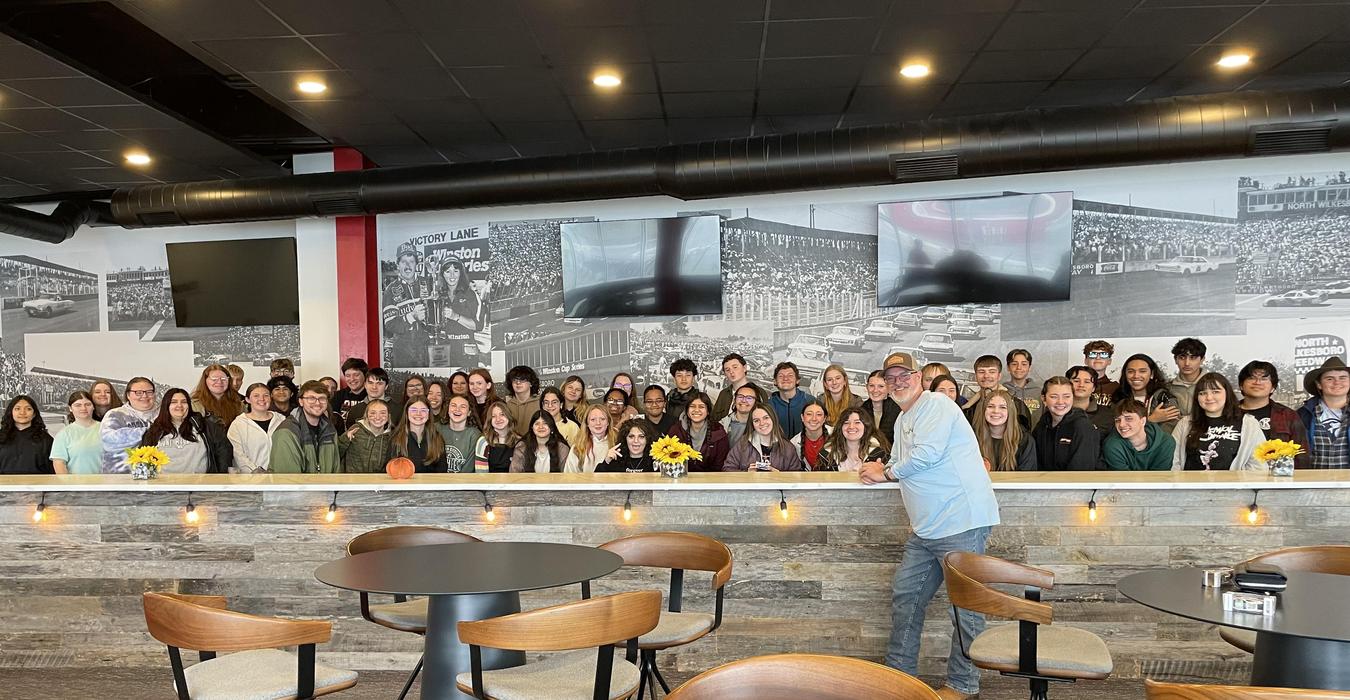 A large group of smiling people gathered in a restaurant with a scenic mural in the background.