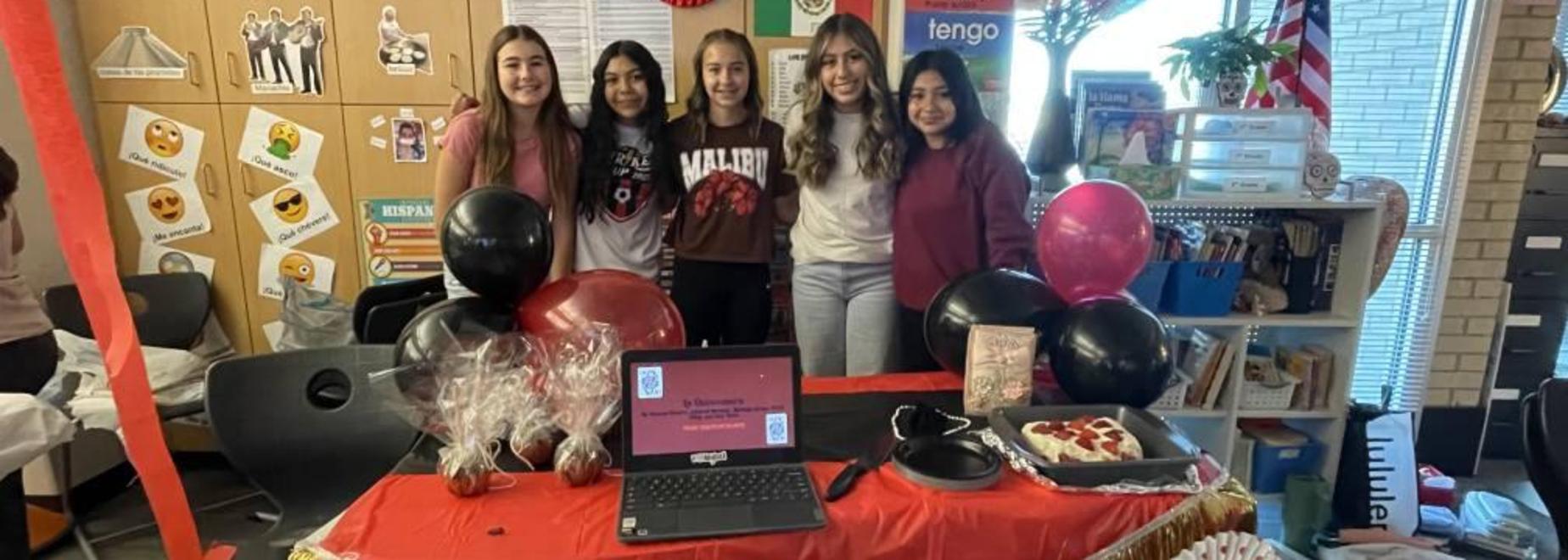 Five girls presenting at a decorated table with a laptop and festive balloons.
