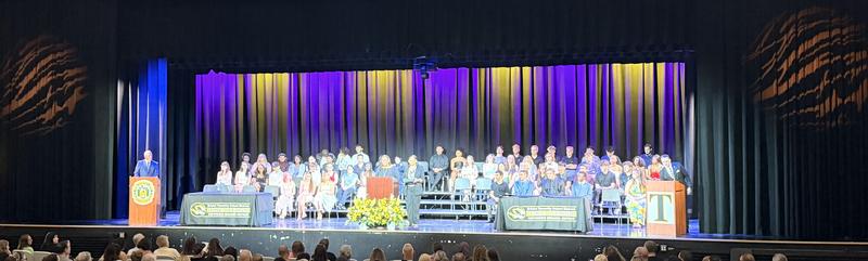 A large group of students sit on an auditorium stage with purple and gold lighting..