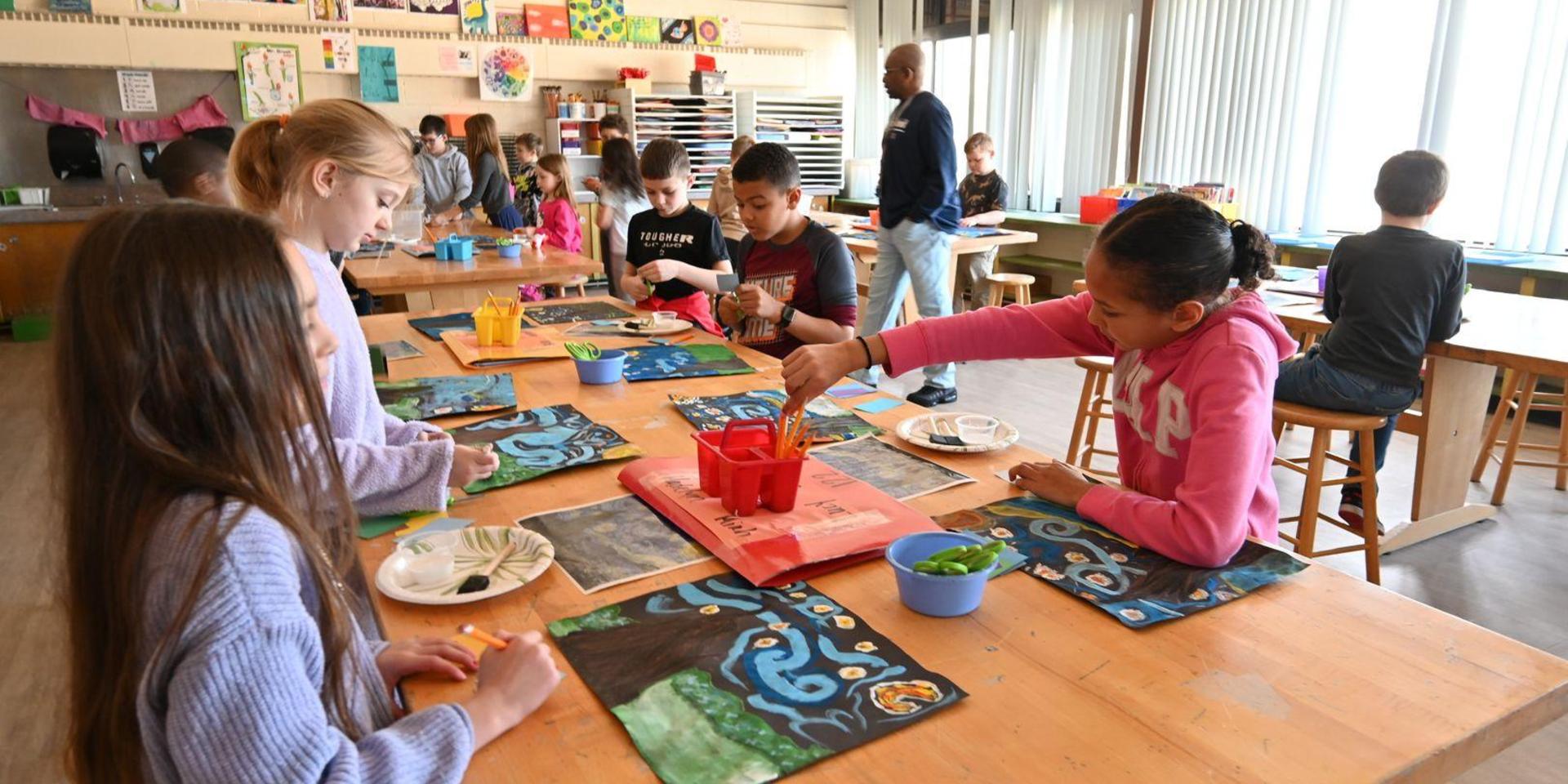 Students creating art at tables with various materials in a bright classroom.