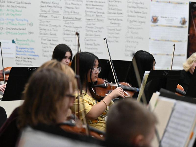 A group of musicians play, with a focus on a girl in glasses playing a violin.