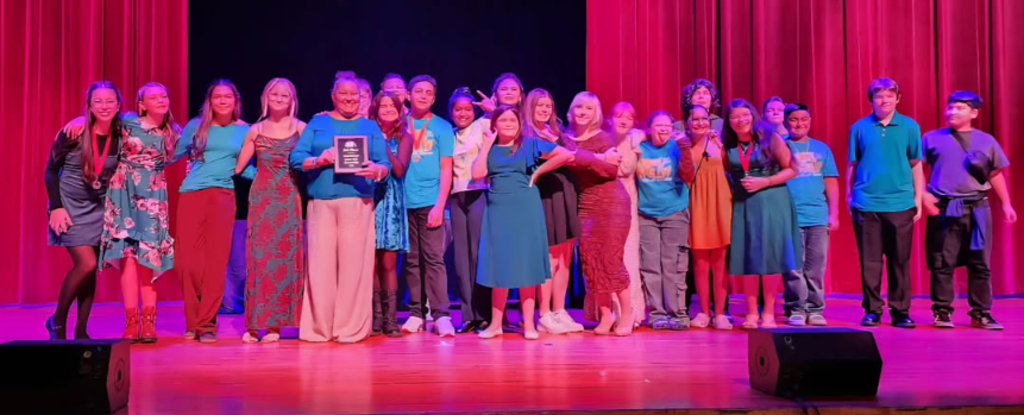 Group photo of individuals celebrating on stage, holding a trophy, in a theater setting.