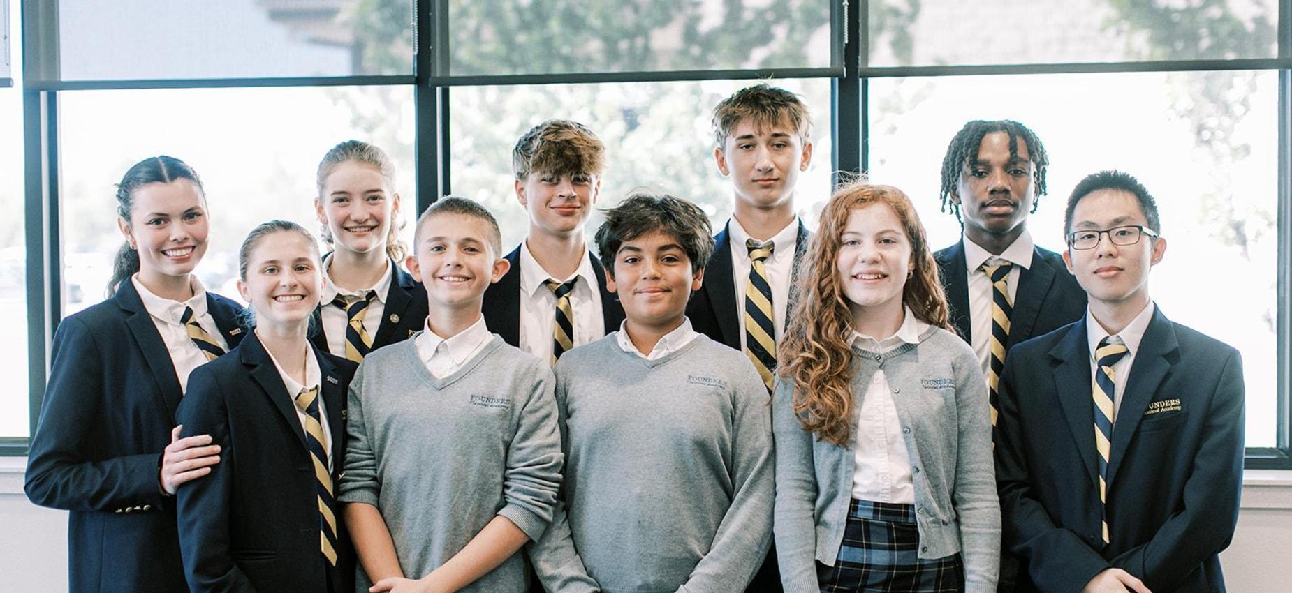Group of students in blazers and sweaters stands together by large windows