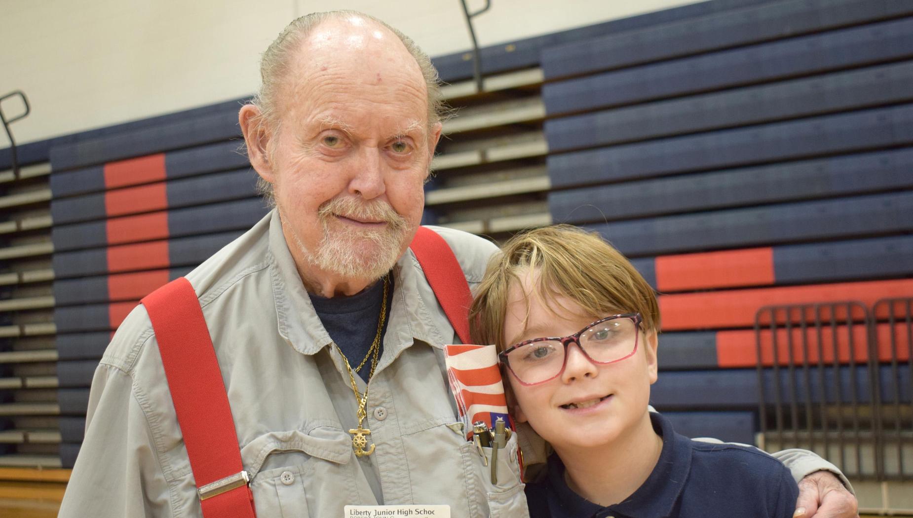 Elderly man with suspenders posing with a boy wearing glasses in a gymnasium.