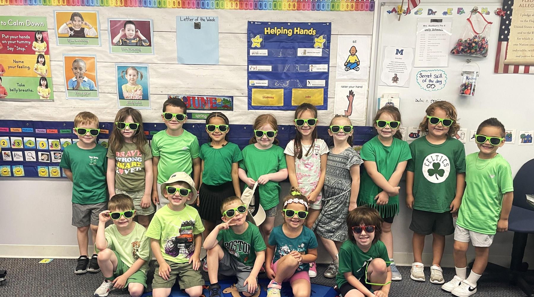 Group of children in green attire wearing sunglasses, posed in a classroom.