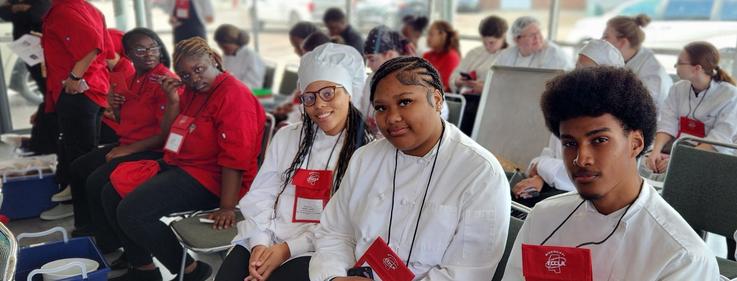 Group of culinary students in chef uniforms seated indoors at an event.