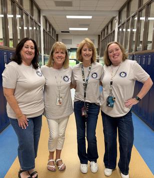 Four women in matching gray t-shirts pose together in a school hallway.