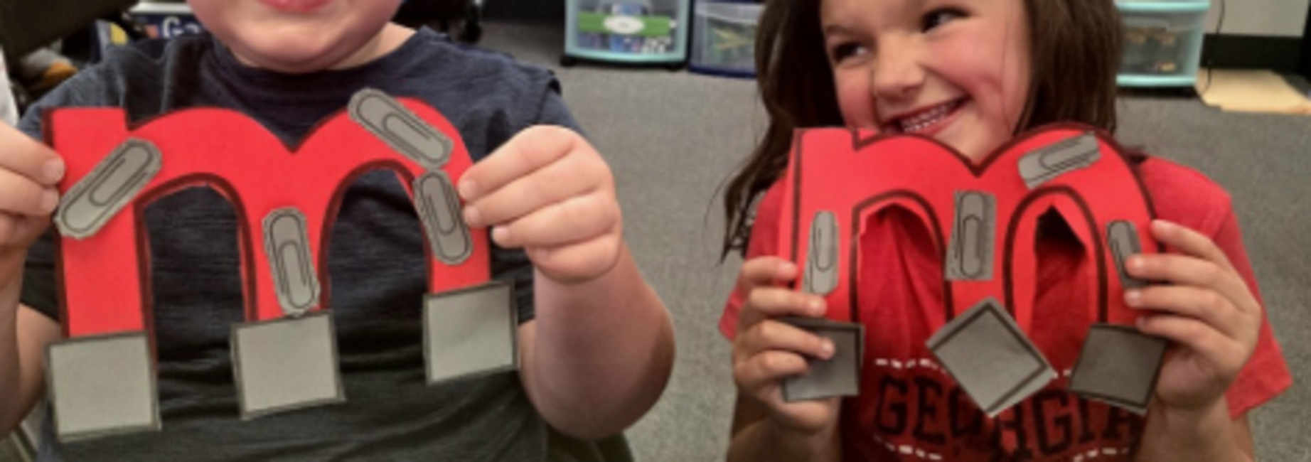 Two children holding large red paper letters 'm' with clips attached.