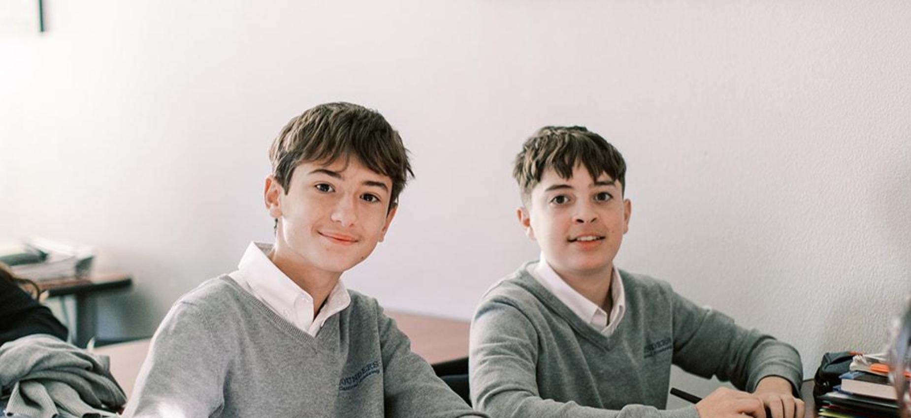 Two smiling students in gray sweaters sit at their desks