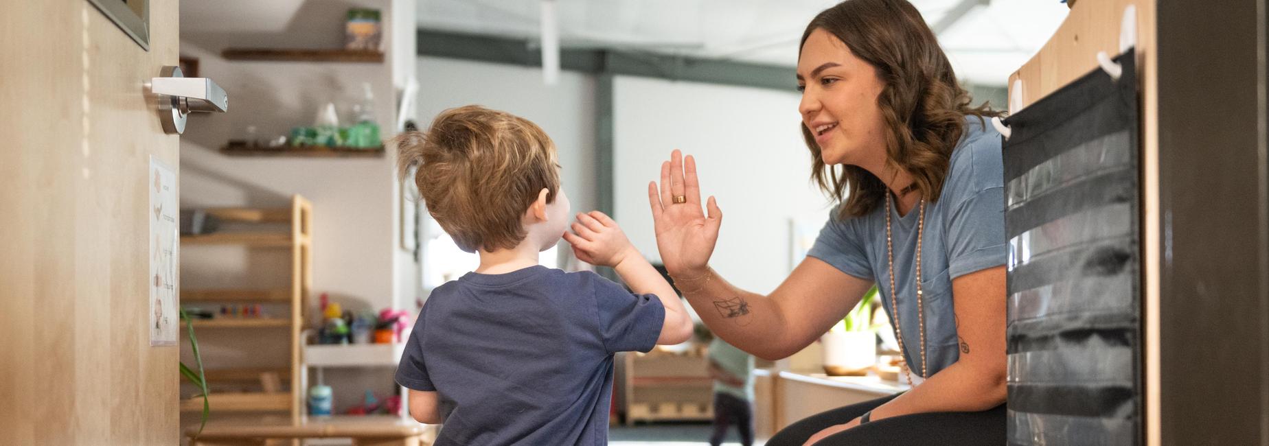 A young boy giving a high five to a smiling adult in a classroom.