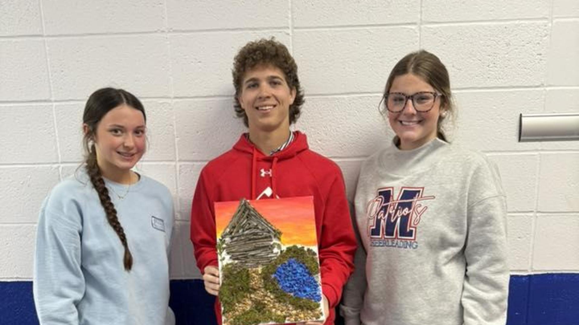 Two students stand beside a classmate holding a colorful painting of a building.
