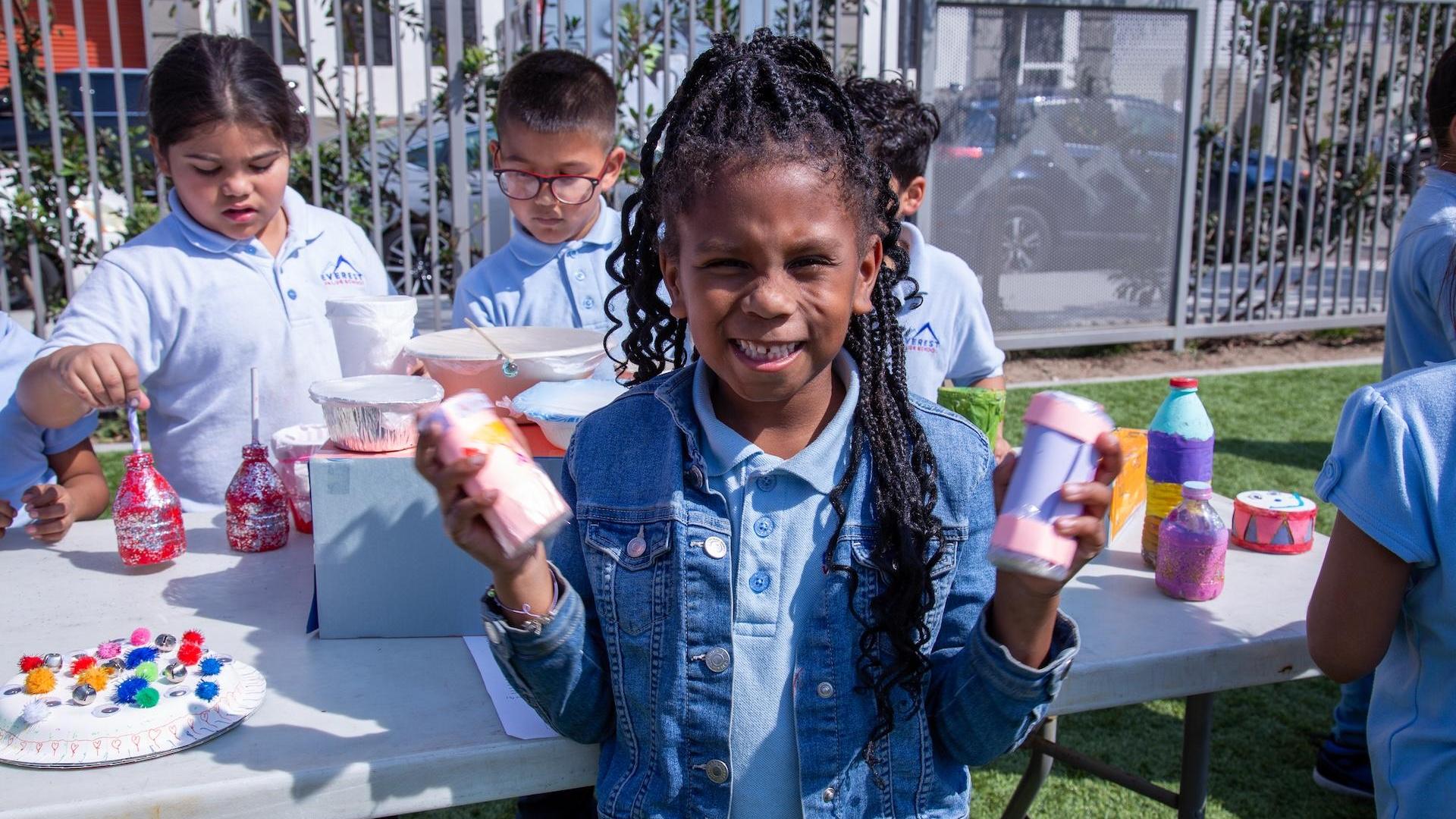 A girl holding painted bottles while surrounded by classmates engaged in art.
