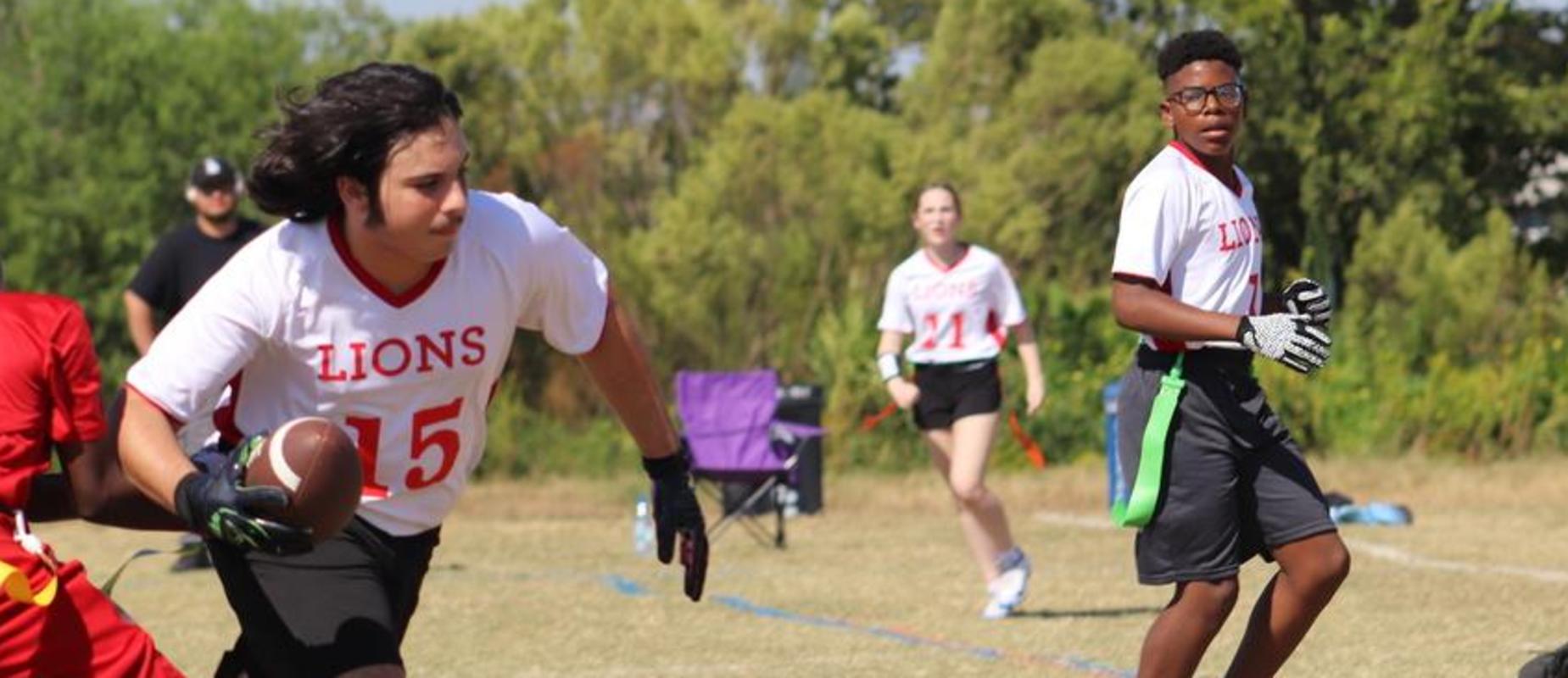 Students run across a field, the one on the left holding a football (from a recent Flag Football game).