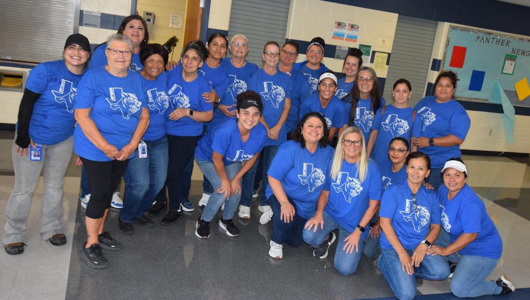 Group of food service employees wearing blue shirts in a school setting, smiling and posing together.