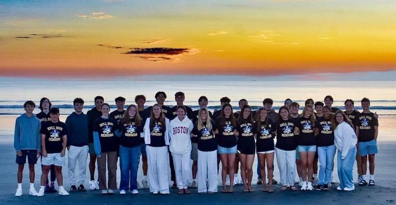Large group of high school students dressed in matching shirts on a beach at sunset.
