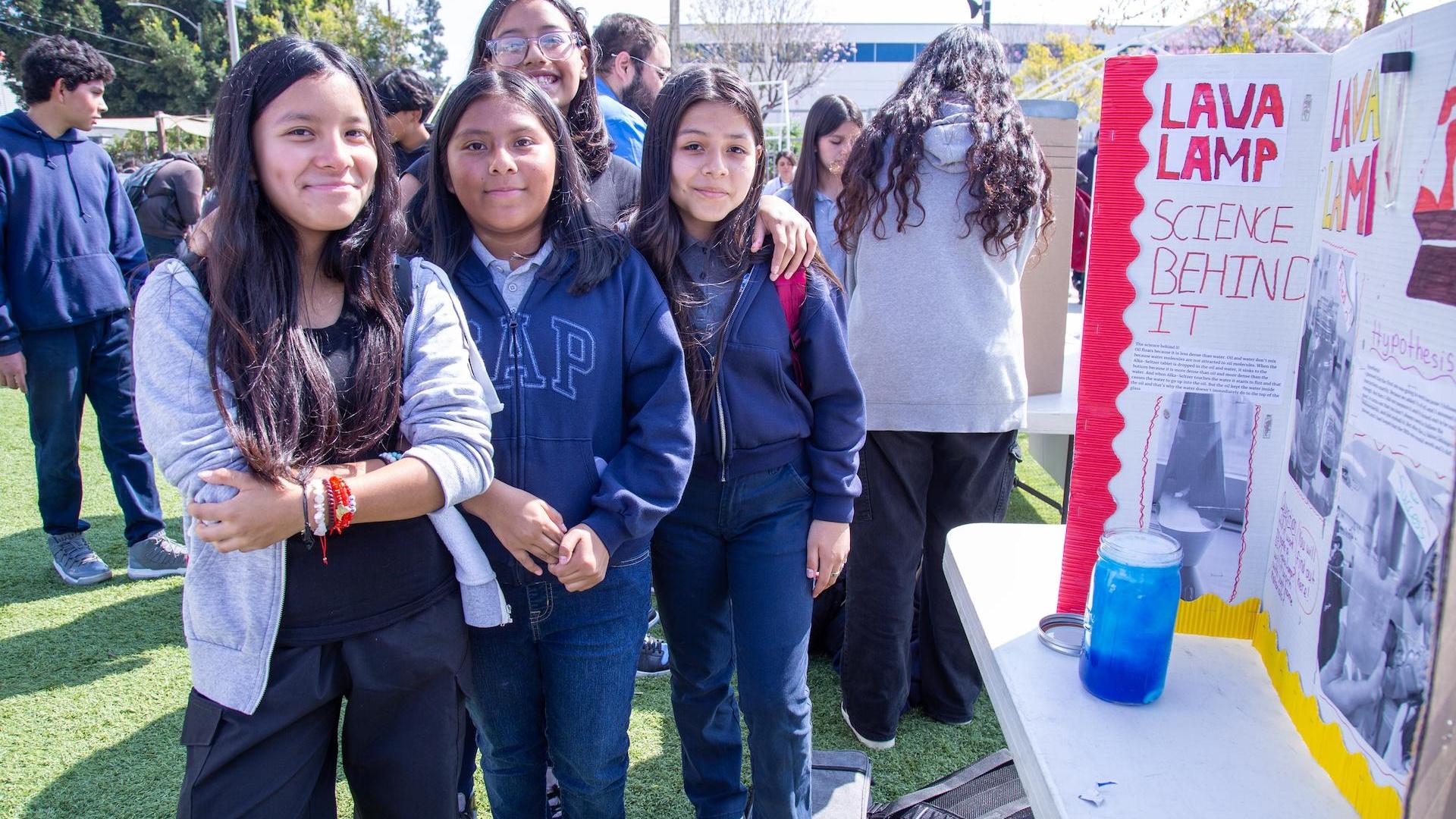 Four girls pose together at a science fair with a project display.