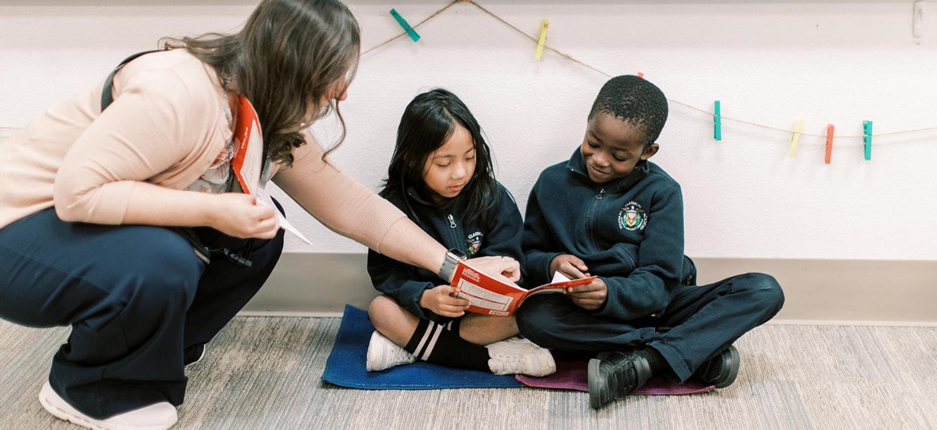 Teacher kneels beside two young students reading on the classroom floor