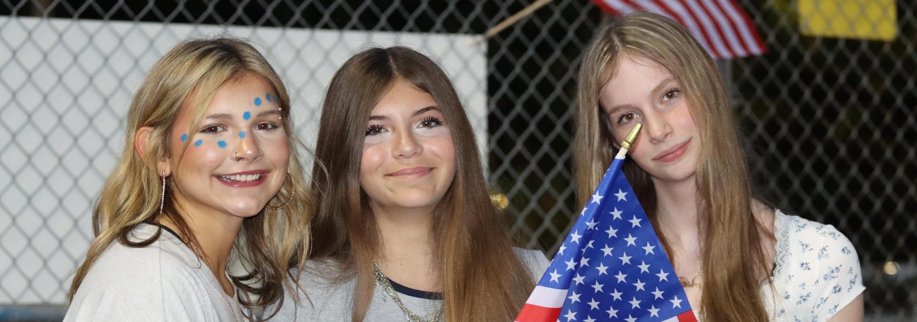 Three young women smiling together, one holding an American flag.