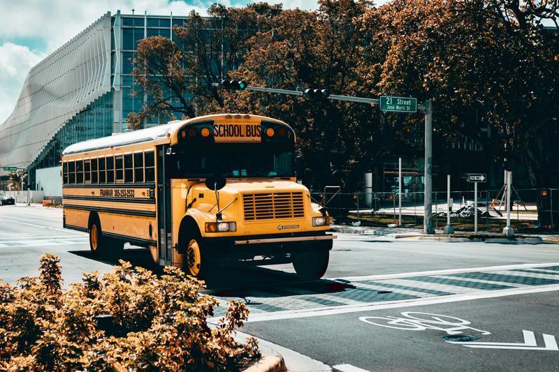 Yellow school bus driving on a city street near trees and modern architecture.