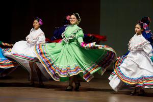 A group of folklorico dancers in colorful traditional dresses perform on stage, mid-spin, with the central dancer in a vibrant green dress smiling brightly.
