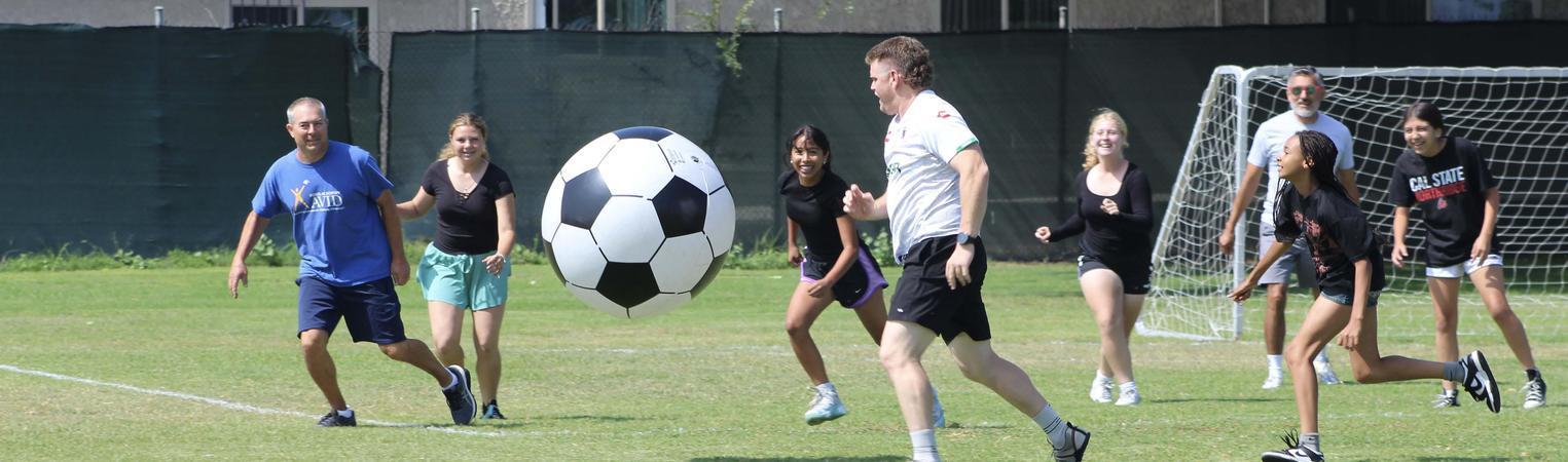Adults and children participate in a soccer game with a large soccer ball.